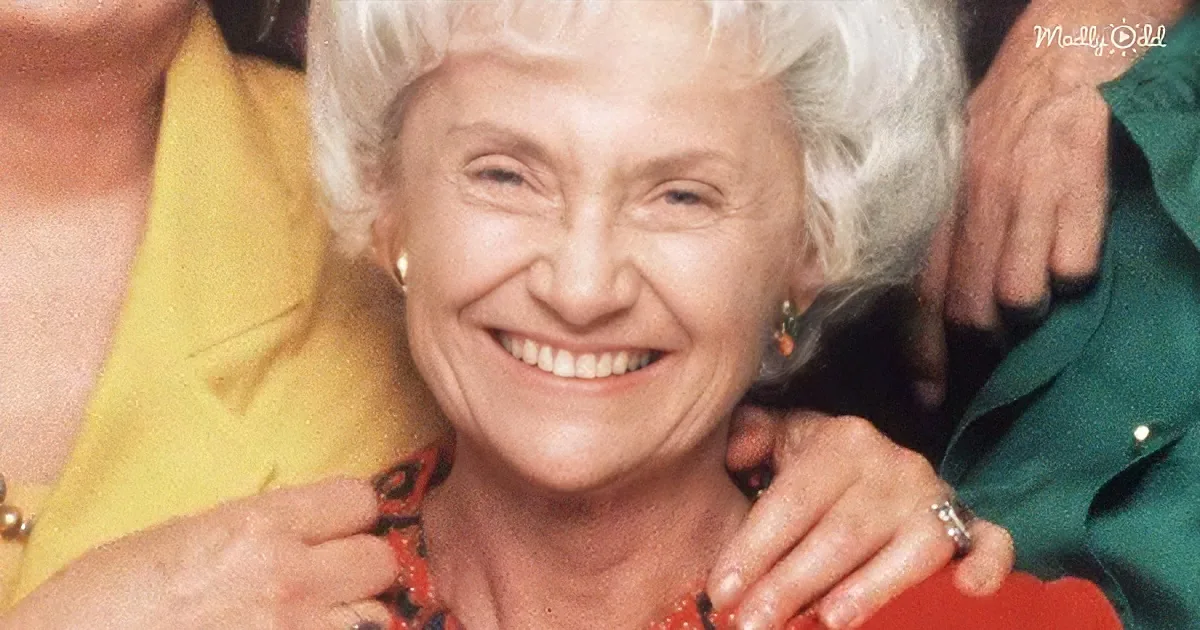 An elderly woman with white hair smiles widely, surrounded by others in a warm embrace.