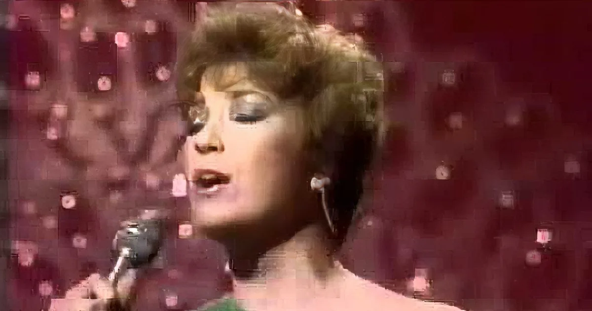 A person with short, curly hair holds a microphone, singing against a red, glittery background.