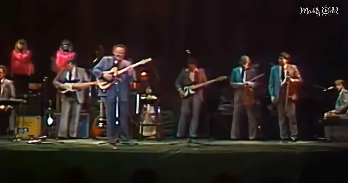 A man in a blue suit plays guitar on stage with a band.