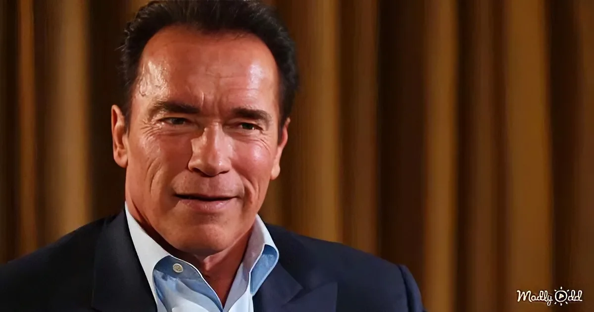 Arnold Schwarzenegger in a suit, smiling, against a golden curtain backdrop.