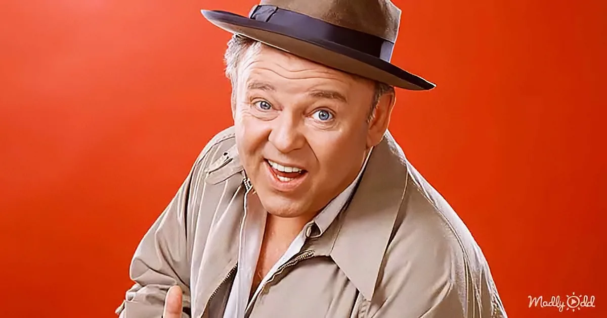 A man in a tan trench coat and fedora hat is smiling and gesturing with his hand.