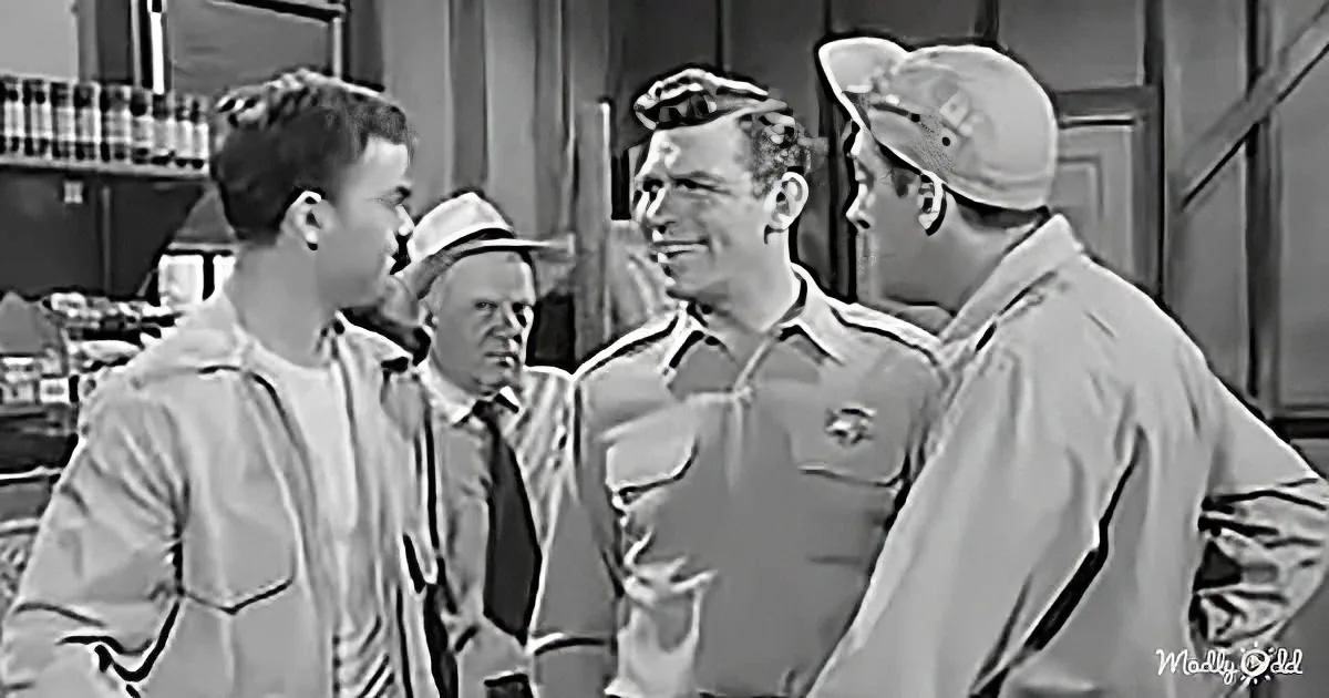 A man in a striped shirt and cap is smiling, surrounded by others in casual attire, in a room with a plaid curtain.