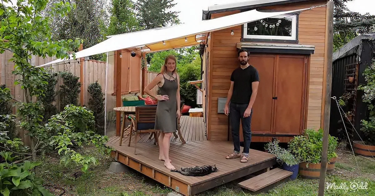 A couple stands on a wooden deck in front of a tiny house with a cat nearby, surrounded by greenery.