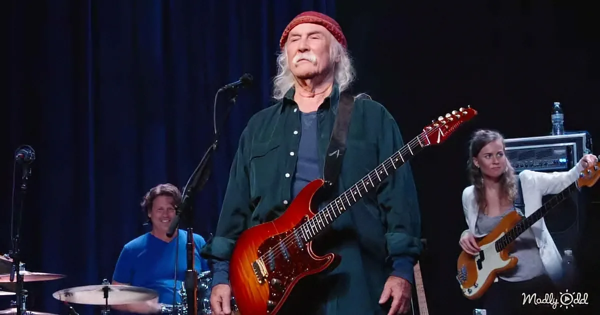 A man in a red beanie plays a red electric guitar on stage with a drummer and another guitarist.