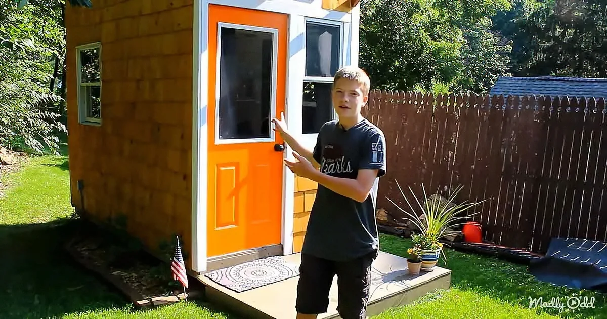A young man stands in front of a small, orange door on a wooden house, gesturing with his hands.