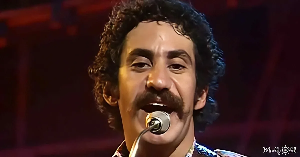 A man with curly hair and a mustache sings into a microphone, wearing a patterned shirt.