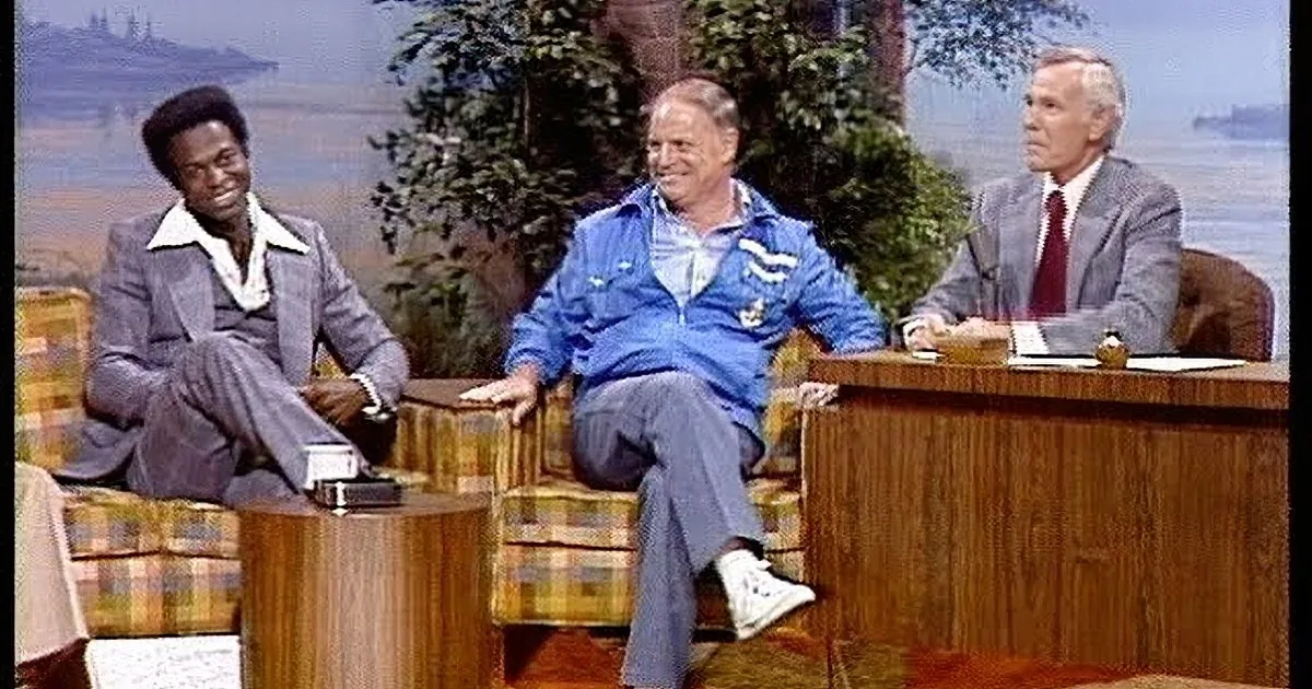 A man in a blue tracksuit is playfully jumping on a couch during a talk show.