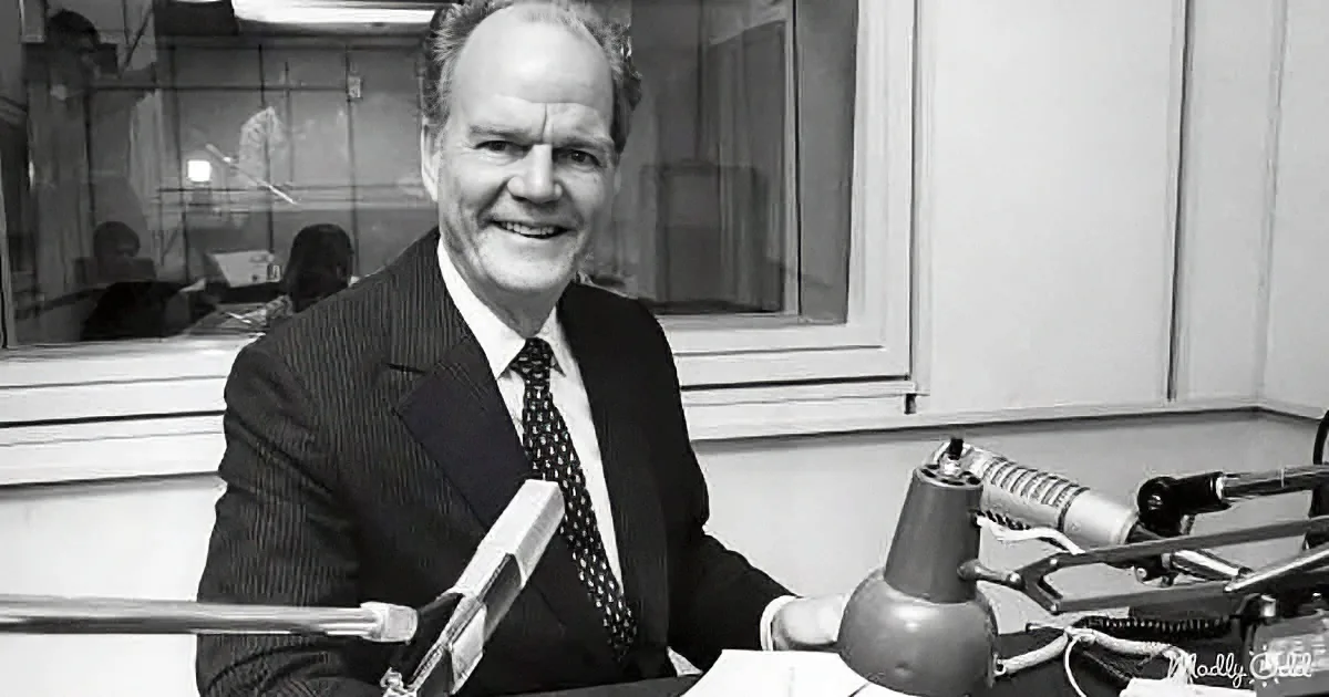 A man in a suit smiles behind a microphone in a radio studio.