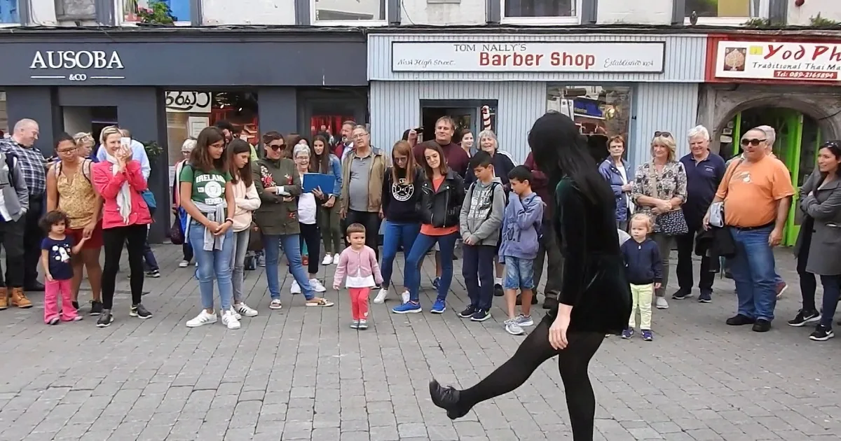 A toddler in a pink sweater and red pants walks on a cobblestone street surrounded by adults.