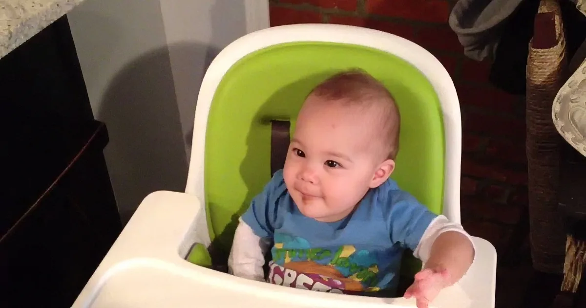 A happy baby in a blue shirt sits in a green and white high chair, smiling broadly.
