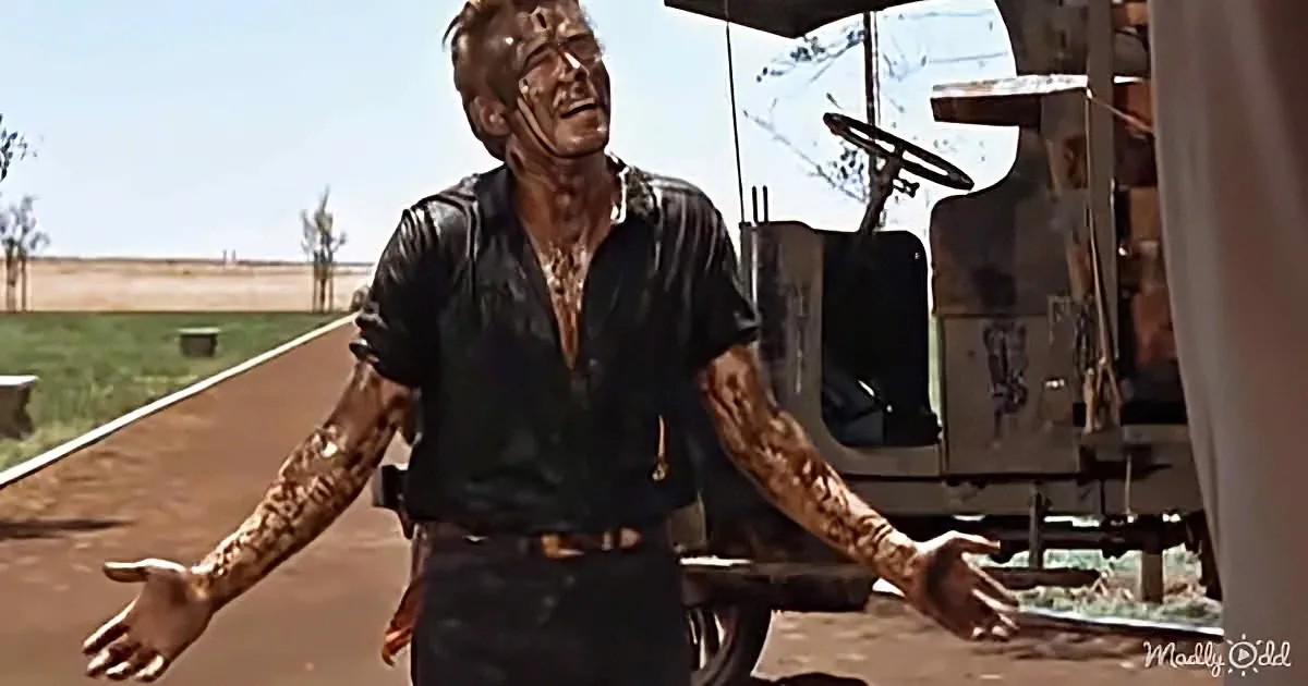 A man covered in mud stands with arms outstretched, looking up, with a truck and open field in the background.