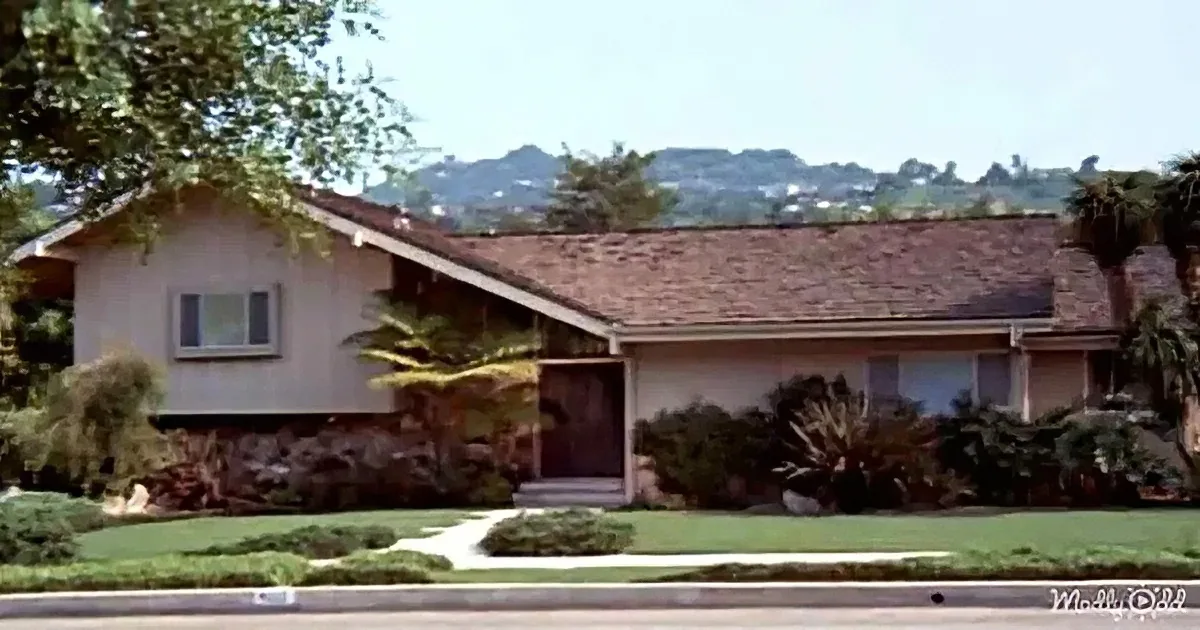 A single-story house with a sloped roof, surrounded by greenery and a clear sky.