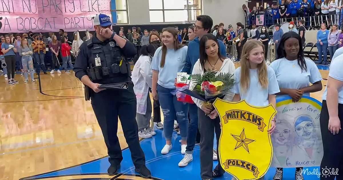 A police officer stands on a basketball court with students, holding a bouquet and a Watkins Police sign.