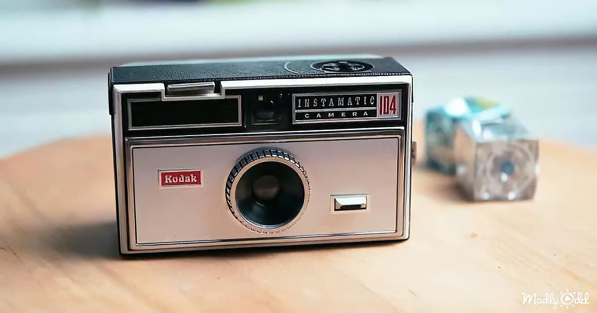 A Kodak Instamatic camera sits on a wooden surface, with a film cartridge in the background.