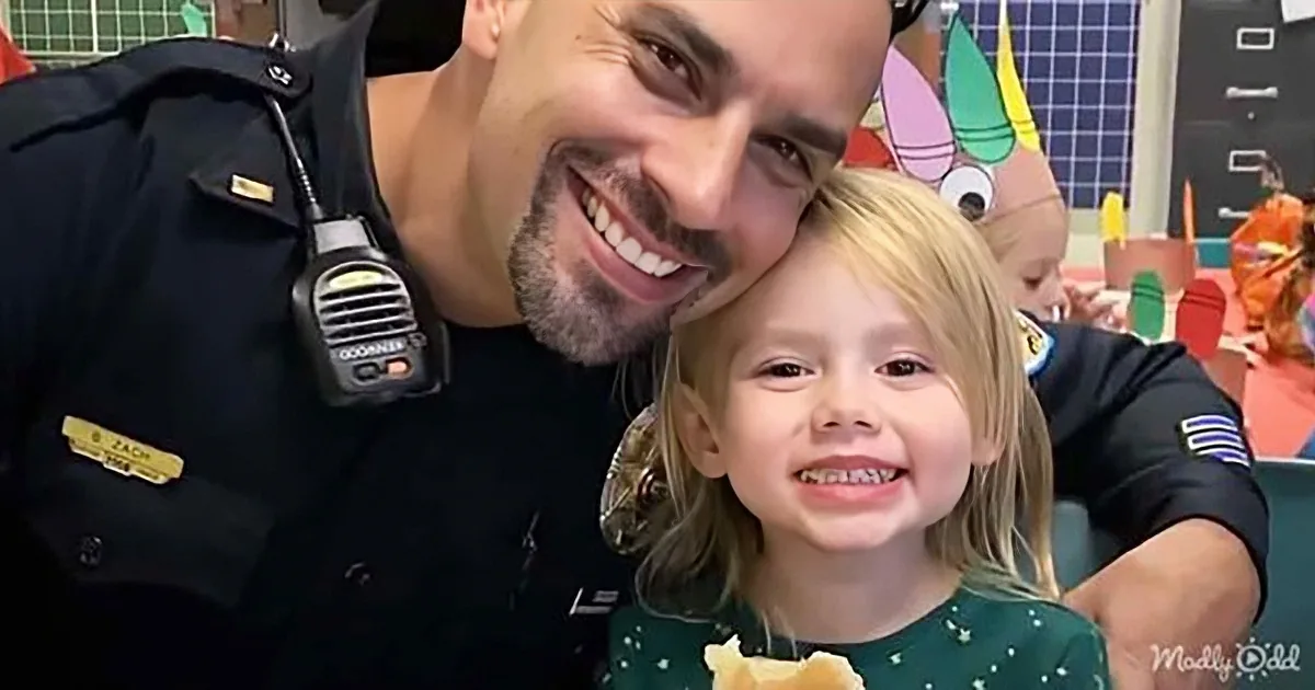 A smiling police officer and a young girl with blonde hair pose together in a classroom setting.