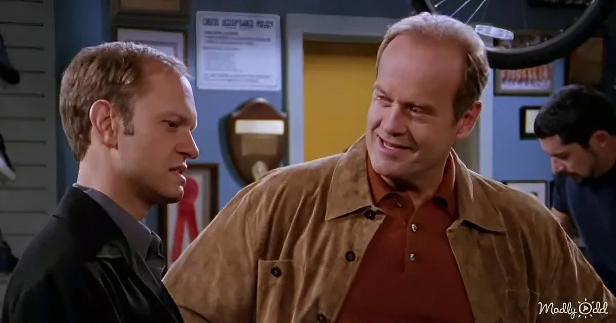 A man in a brown jacket and orange shirt is smiling and talking to another man in a black jacket in a room with a yellow...