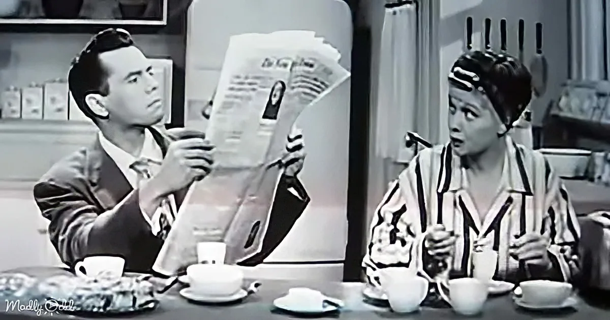 A man reads a newspaper while a woman sits at a table, both holding cups, in a vintage kitchen setting.
