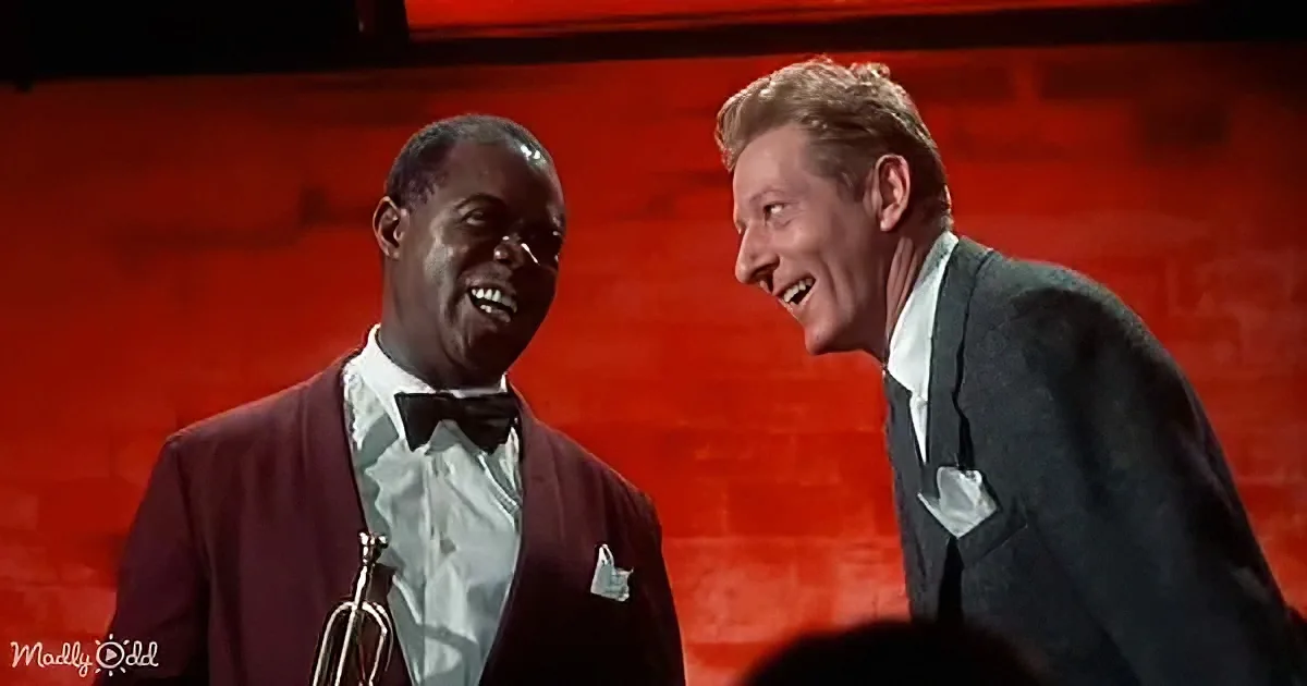 Two men in suits smile and laugh, one holding a trumpet, against a red background.