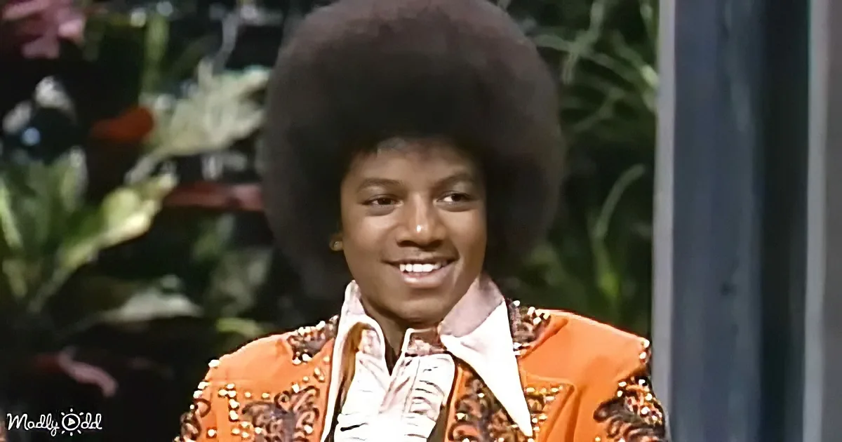 A smiling person with a large afro hairstyle, wearing an orange jacket with studs, stands in front of a green, leafy...