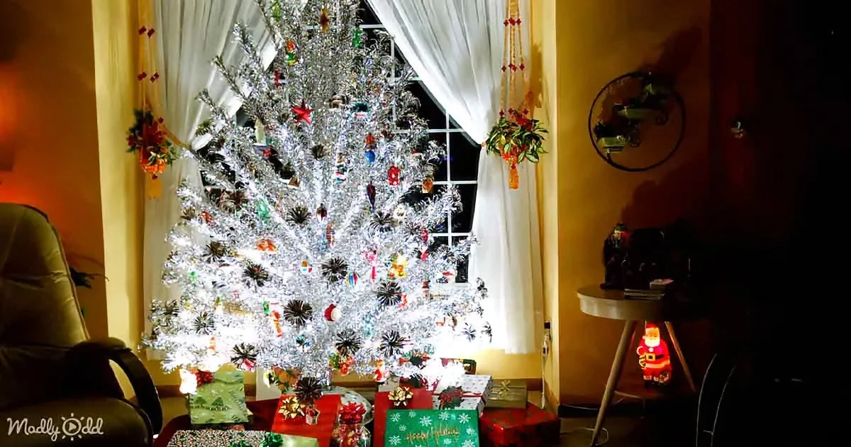 A beautifully decorated silver Christmas tree with ornaments and lights stands in front of a window, surrounded by...