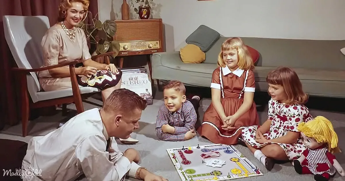 A family is gathered around a board game on the floor, with a woman in a chair and a man in a white shirt.