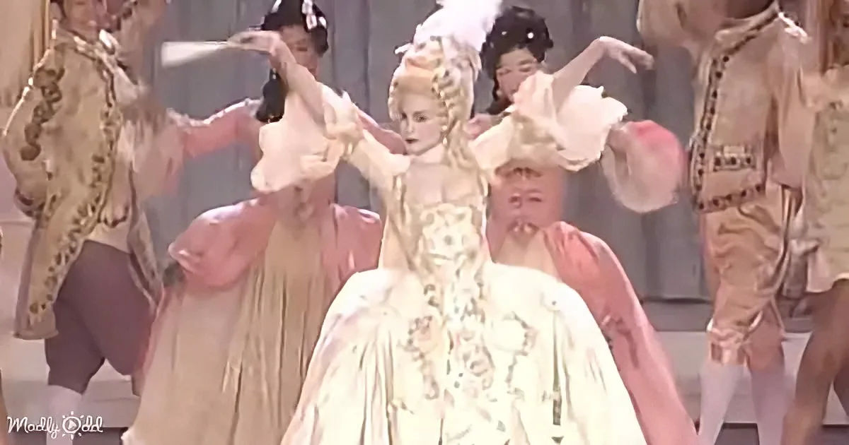 A woman in a white, ornate gown with a large, elaborate headdress is surrounded by dancers in period costumes.