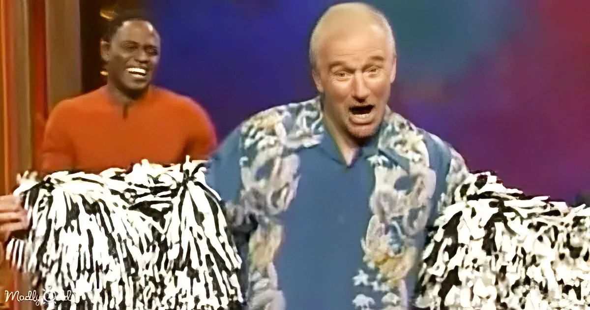 A man in a floral shirt holds pom-poms, appearing surprised, with another man in an orange shirt smiling behind him.
