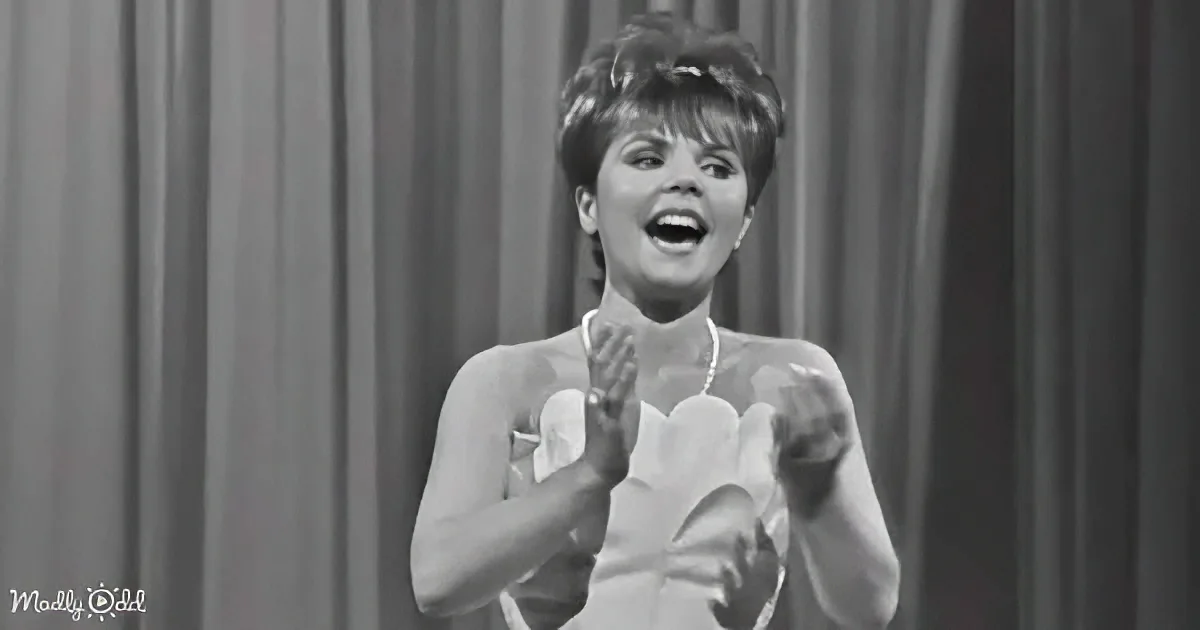 A woman in a white dress claps her hands, smiling, against a curtain backdrop.