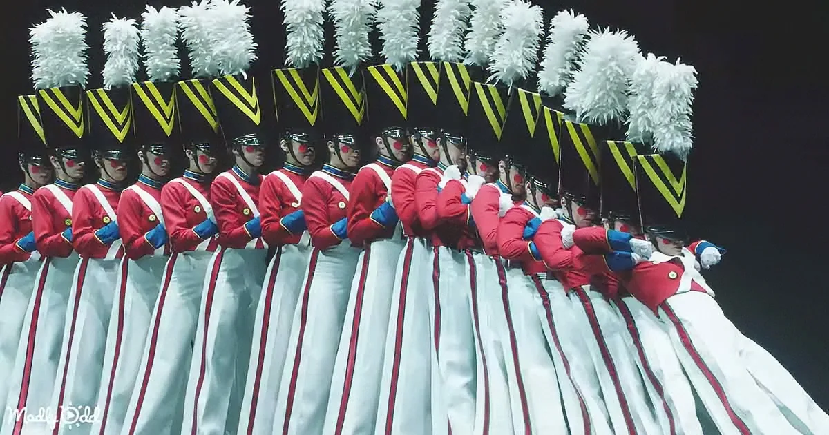 A group of uniformed performers in red jackets and white pants with tall hats and white plumes.