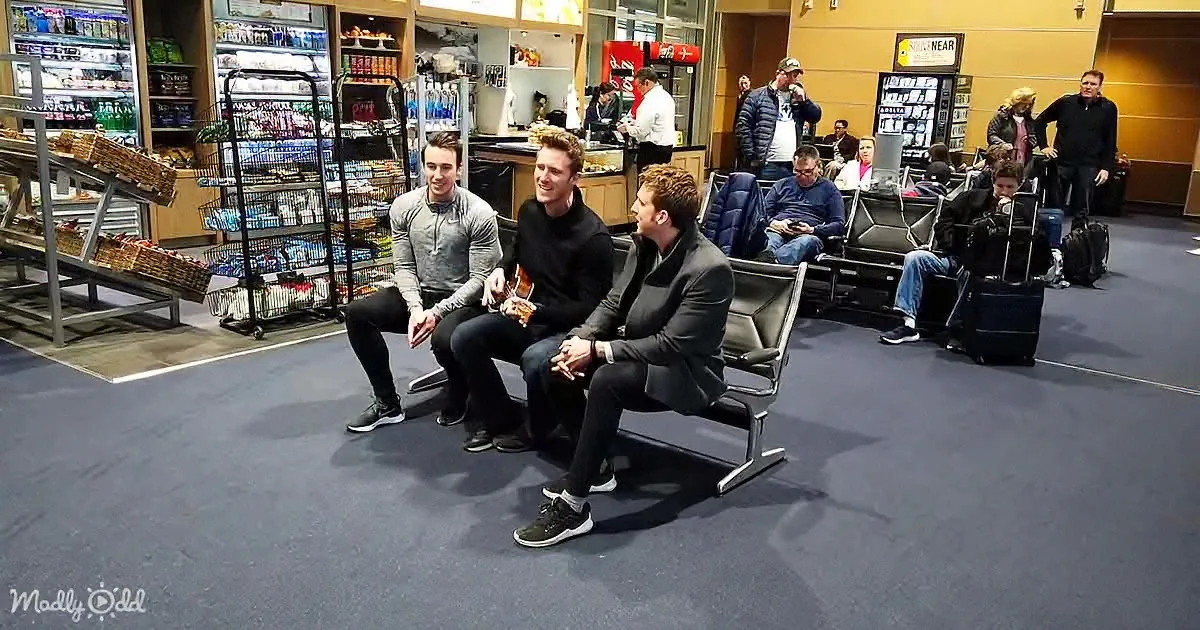 Four men sit on a bench in an airport lounge, smiling and talking, with a store and other travelers in the background.