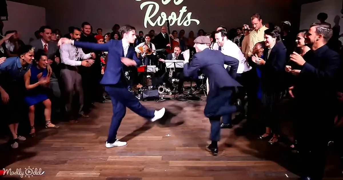 Two men in suits dance energetically on a wooden floor surrounded by a cheering crowd and a band.