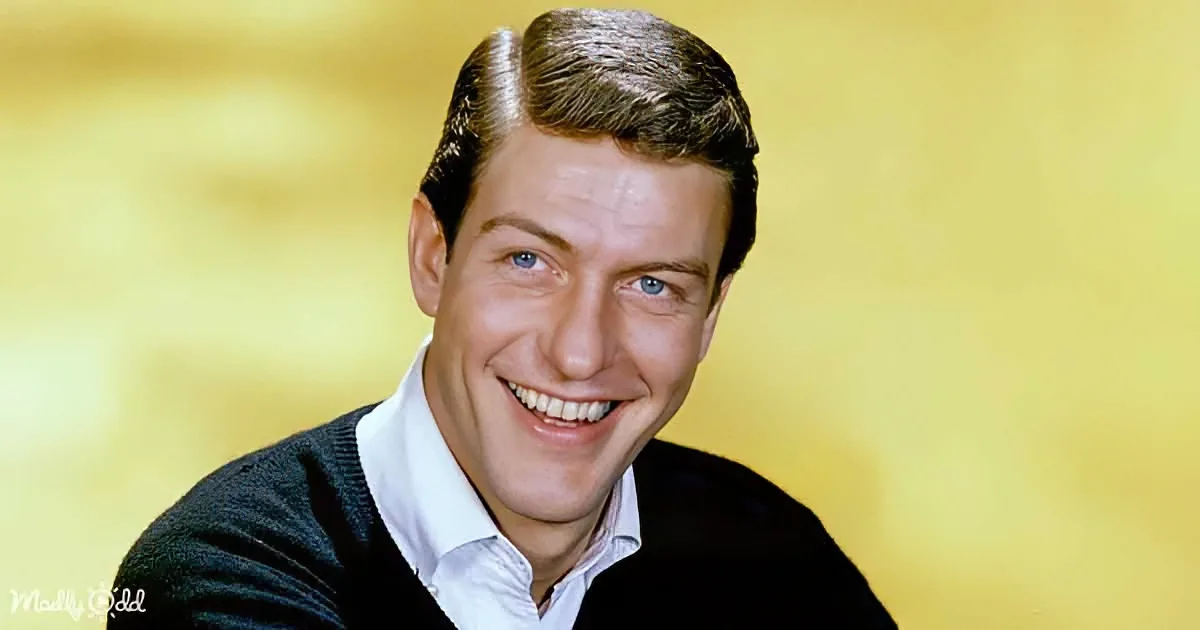 A smiling man with short brown hair, wearing a black sweater over a white shirt, against a yellow background.