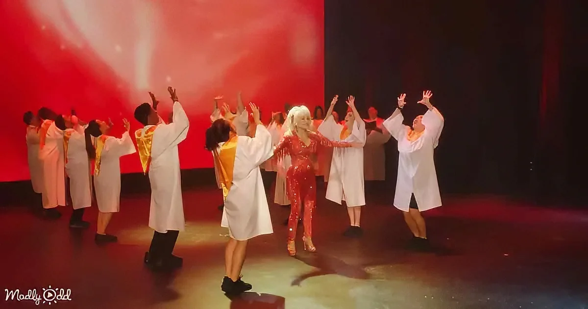 A performer in a red sequined outfit dances on stage with a group in white robes, under a red backdrop.
