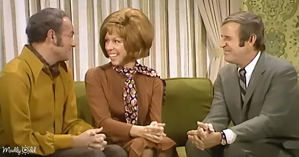 Three people sit on a green couch, two men and a woman in a floral scarf, smiling and engaged in conversation.