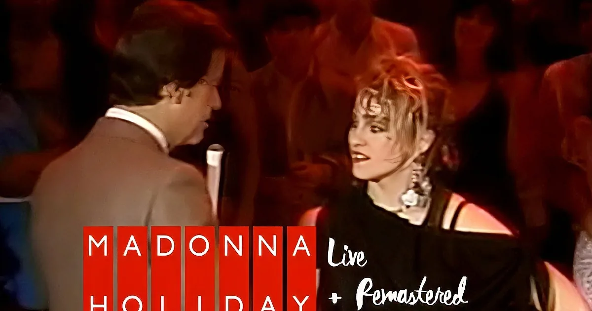 A man and woman are singing on stage, both holding microphones, with a crowd in the background.