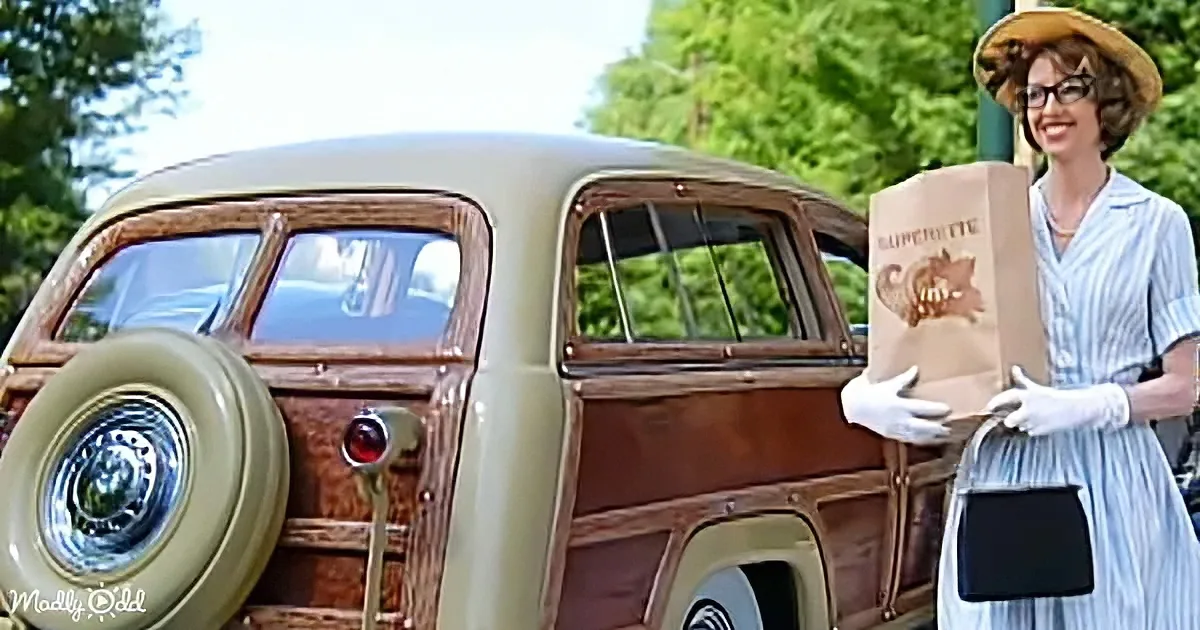 A woman in a vintage dress holds a paper bag near a classic car with a wooden panel design.