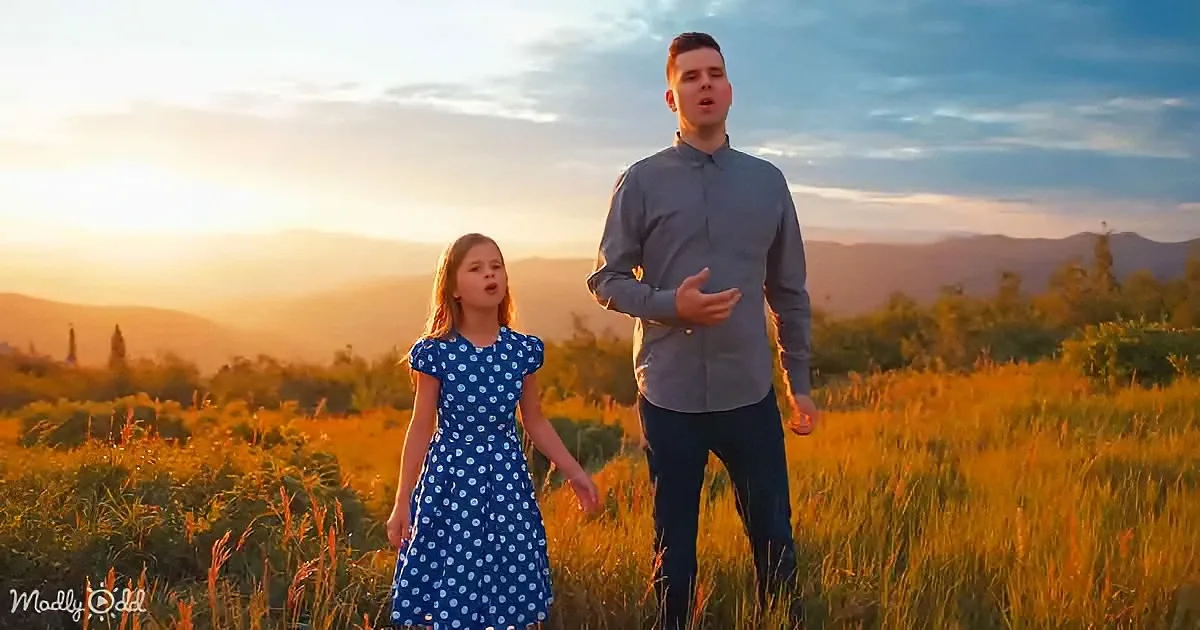 A man and a young girl sing in a field at sunset, surrounded by golden grass and distant hills.