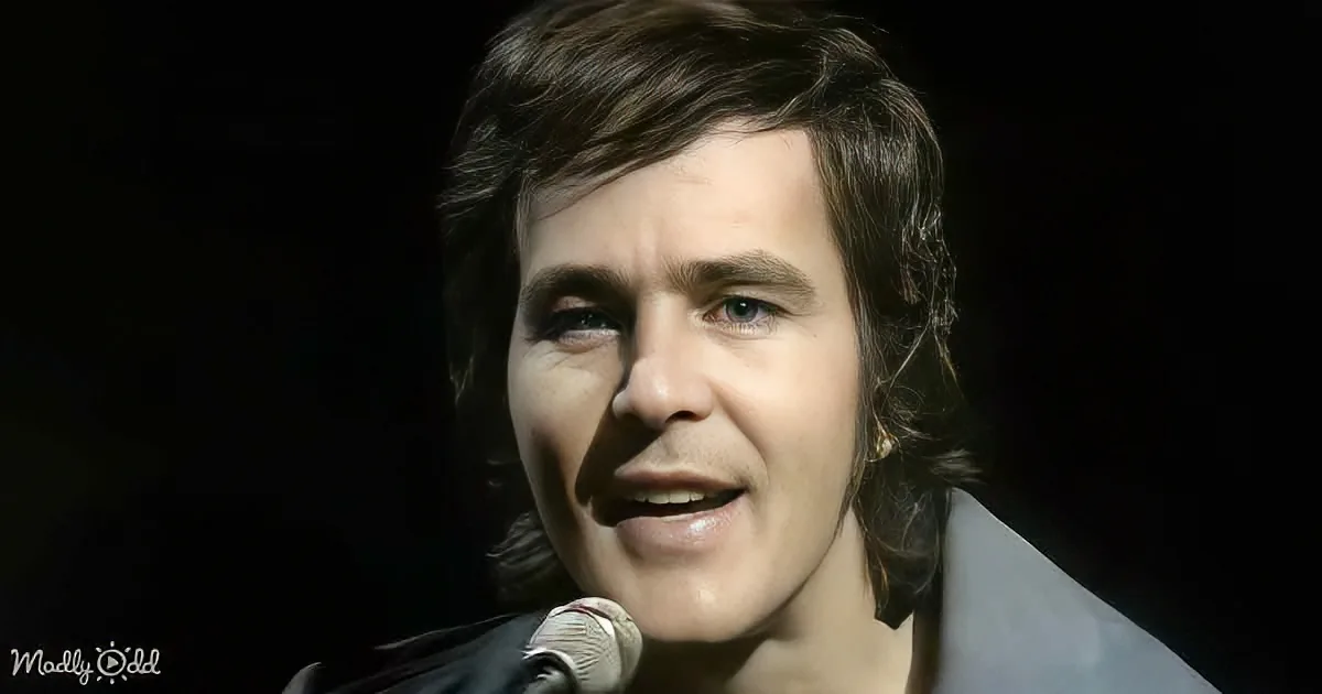 A person with shoulder-length hair smiles near a microphone against a dark background.
