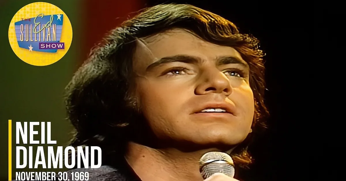 A man with dark, wavy hair and a microphone, possibly singing or speaking, in a dimly lit setting.