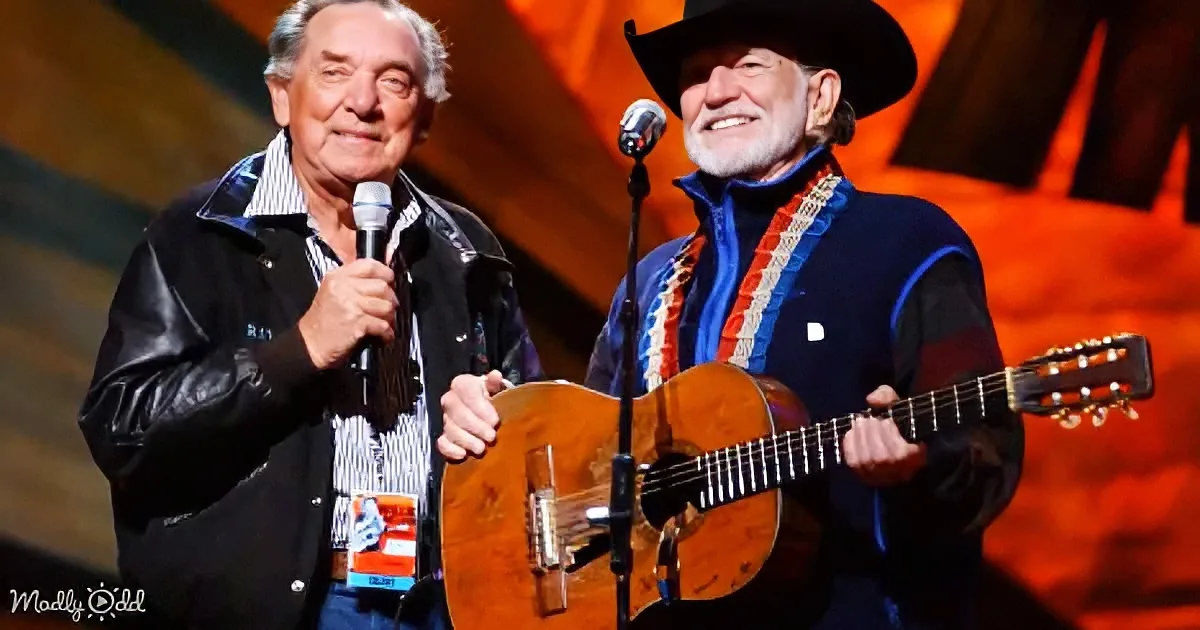 Two men on stage, one holding a microphone, the other playing guitar, both smiling.