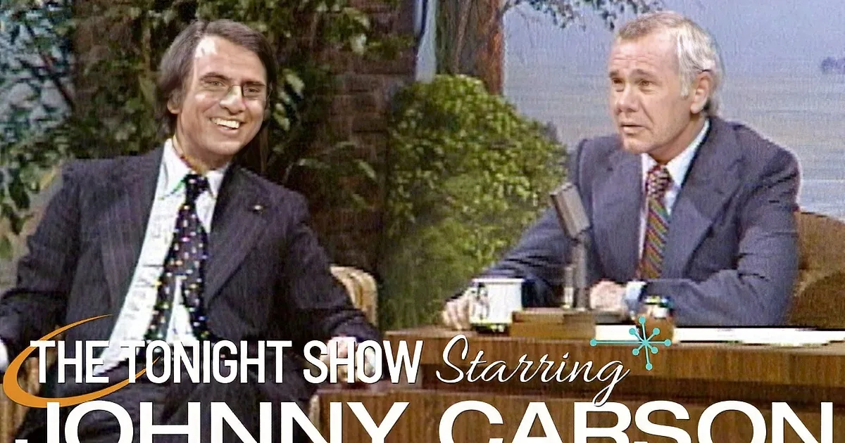 Two men in suits smile on a talk show set with a wooden desk and plants in the background.
