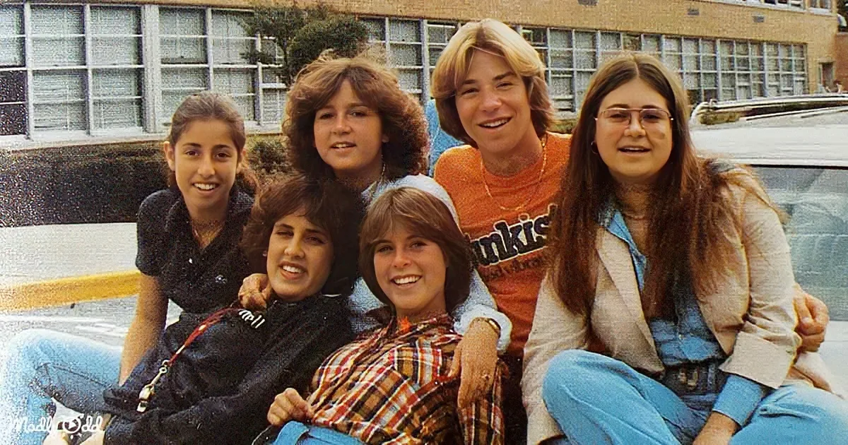 Six young people, including a boy in an orange shirt, are sitting together outdoors, smiling and posing for a photo.