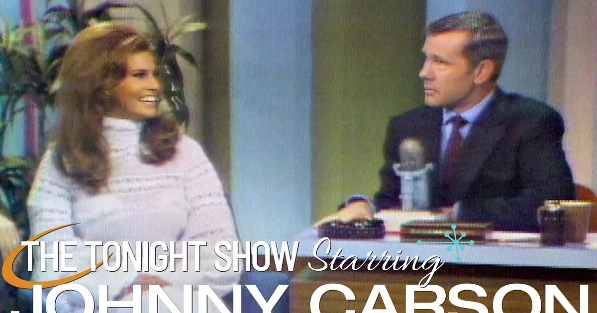 A woman in a white dress smiles next to a man in a suit at a desk with a microphone.