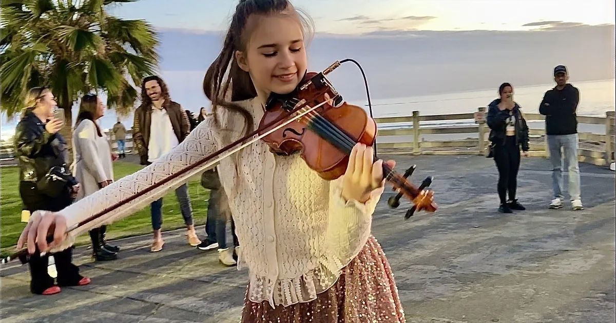 A girl in a white dress plays the violin on a pier at sunset, with a person in a red dress walking in the background.