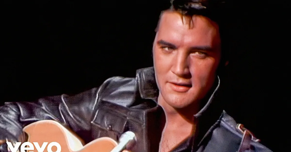 A person in a leather jacket plays a guitar and sings, with a dark background.