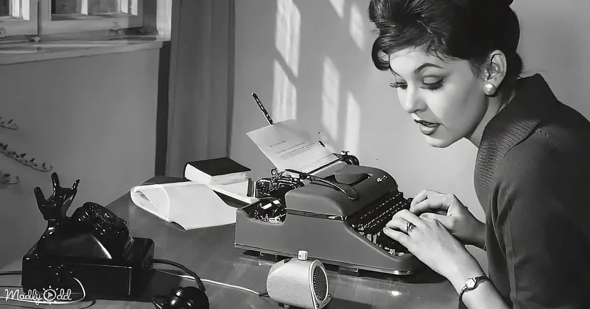 A woman types on a vintage typewriter, surrounded by books and a telephone, in a well-lit room.