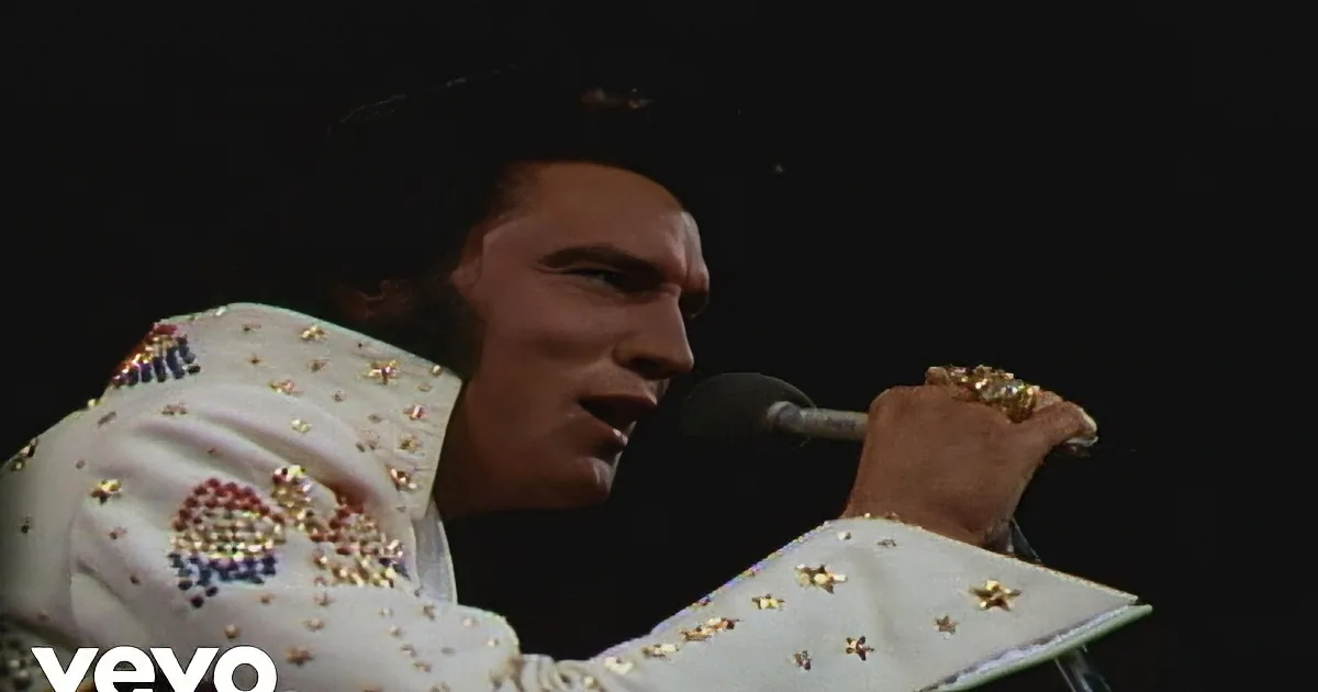 A person in a white star-adorned suit sings into a microphone, holding a gold ring.