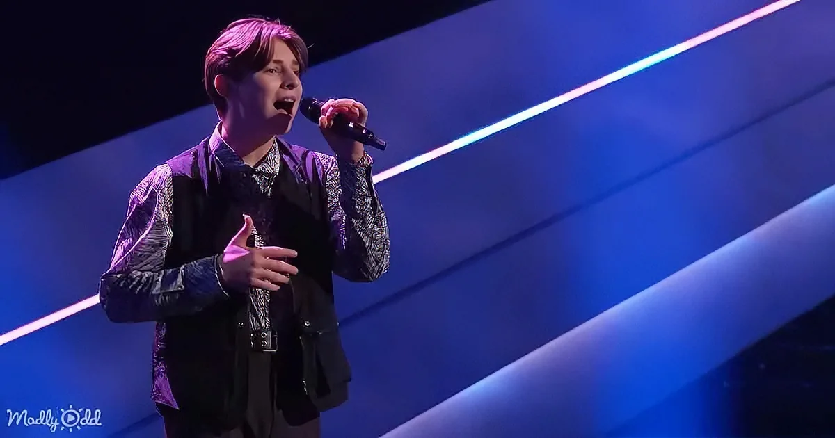 A person in a purple and black outfit sings into a microphone on a stage with blue lighting.