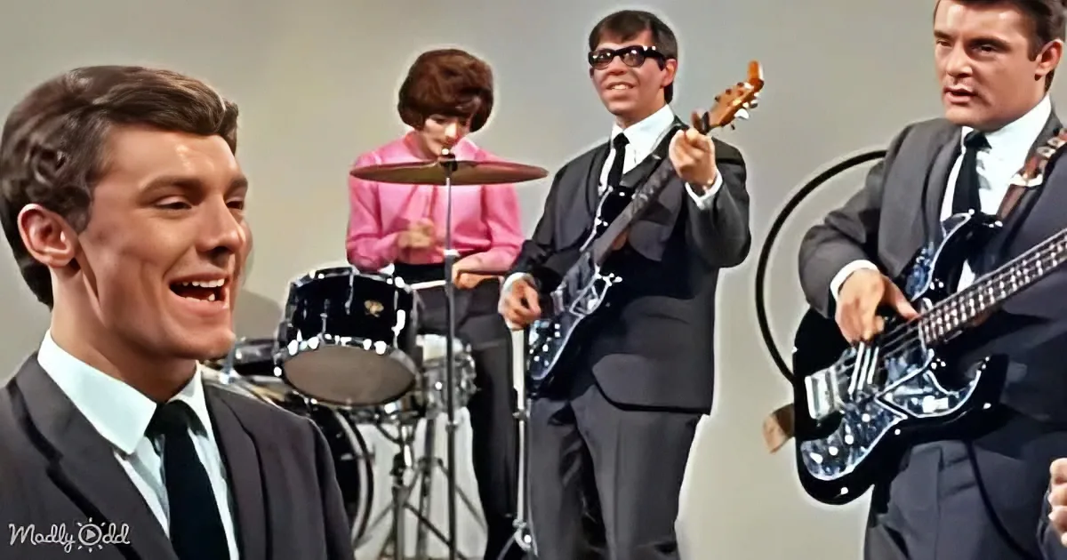 A smiling man in a suit and tie leads a band playing guitars and drums.