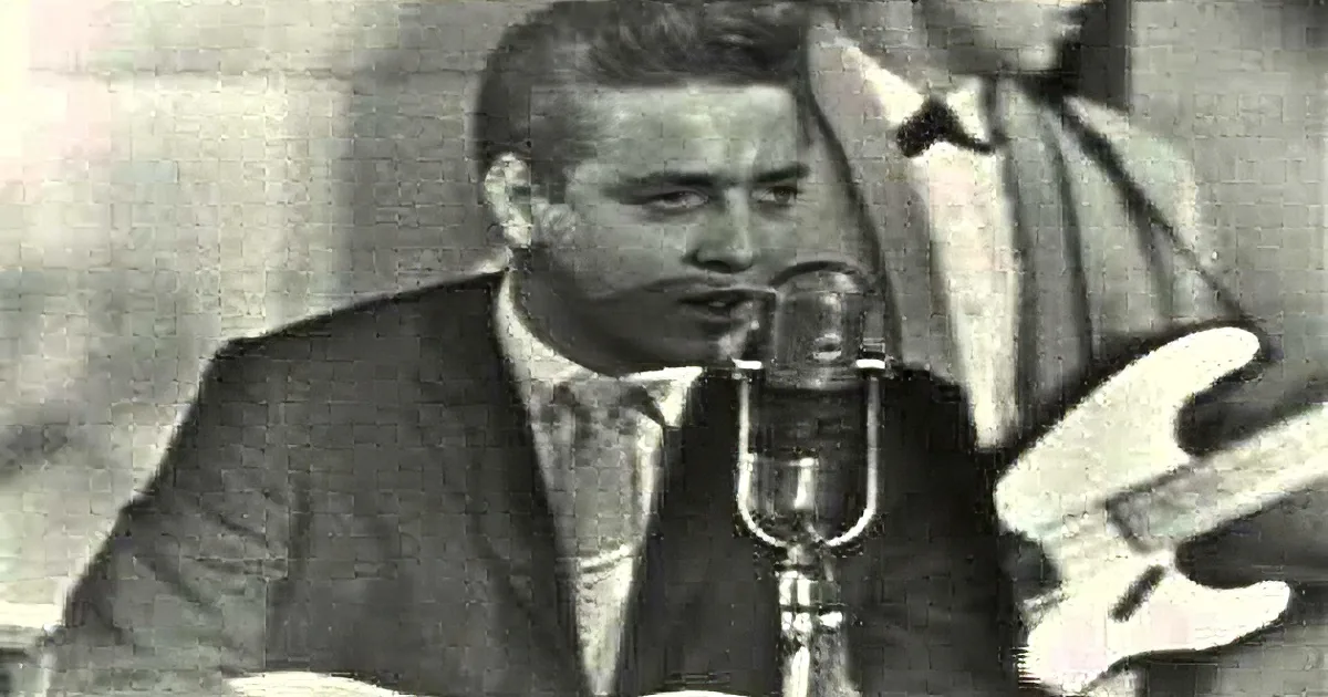 A man in a suit sings into a microphone, with another person playing a guitar in the background.